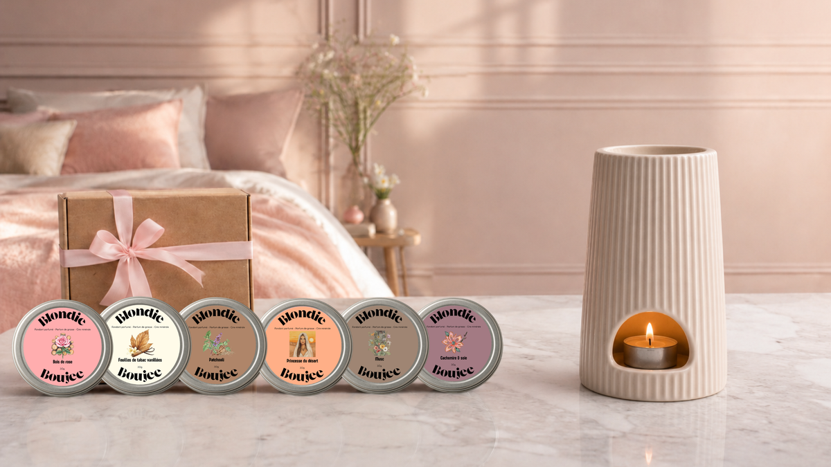 Un brûle parfum avec une bougie chauffe plat allumée sur une table en marbre blanche avec à côté coffret de fondants parfumés Blondie Boujee